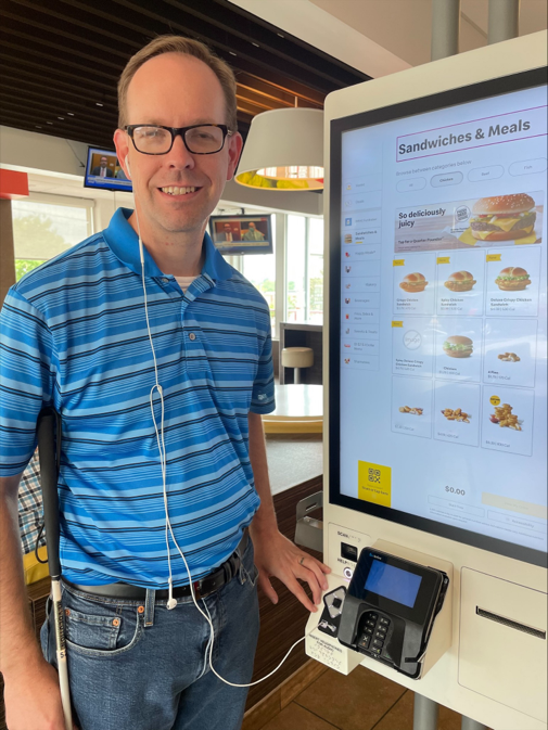 A customer holding a cane with ear buds, smiling and placing a food order using an accessible self-service kiosk at a restaurant.